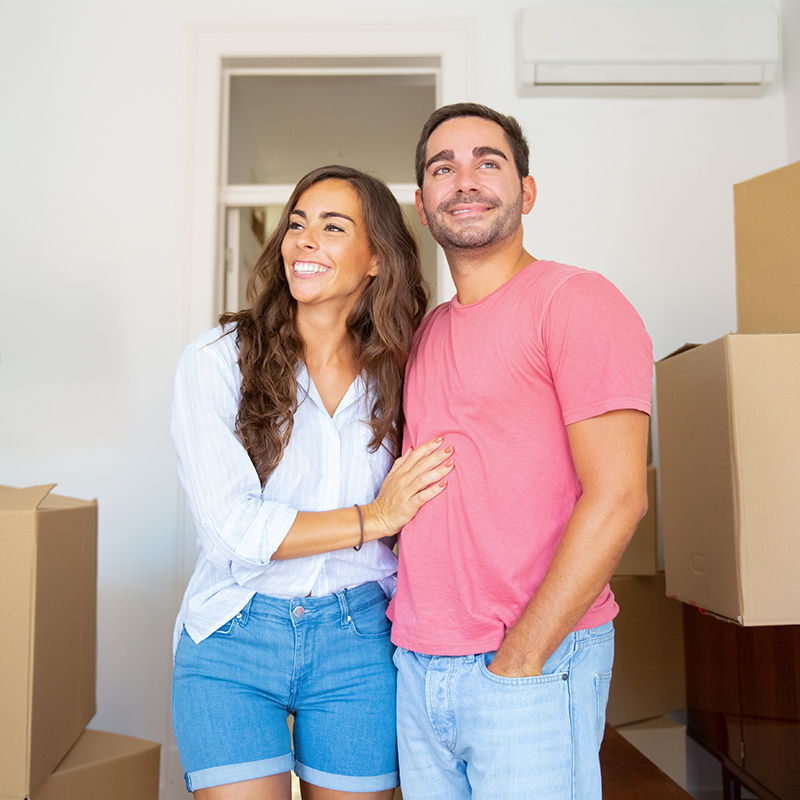 Casal sorridente abraçado em nova casa com caixas de mudança ao fundo.