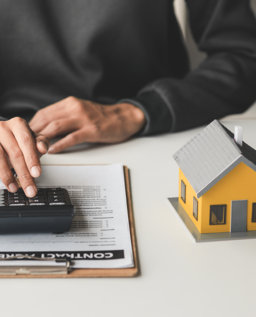 Simulação de crédito habitação com calculadora e contrato ao lado de maquete de casa, representando as despesas associadas à compra de imóvel.
